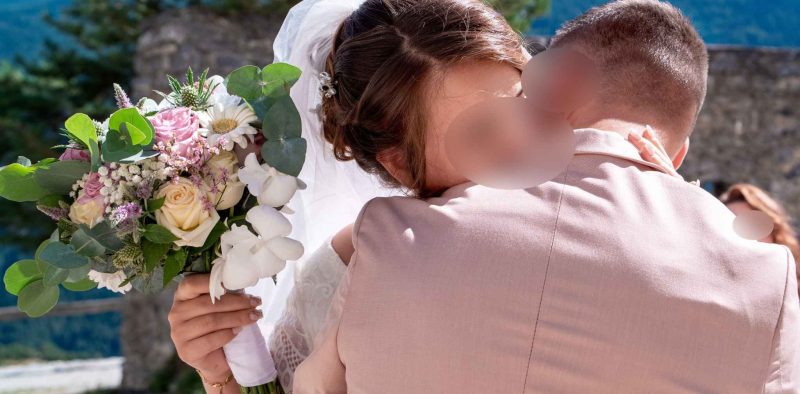 Une mariée enlace son époux en tenant un bouquet fleuri, avec en arrière-plan les montagnes et la nature.