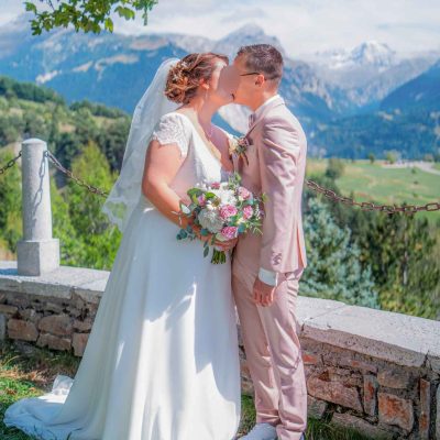 Les mariés s’embrassent tendrement face à un panorama de montagnes enneigées, par une journée ensoleillée. La mariée tient un bouquet fleuri.