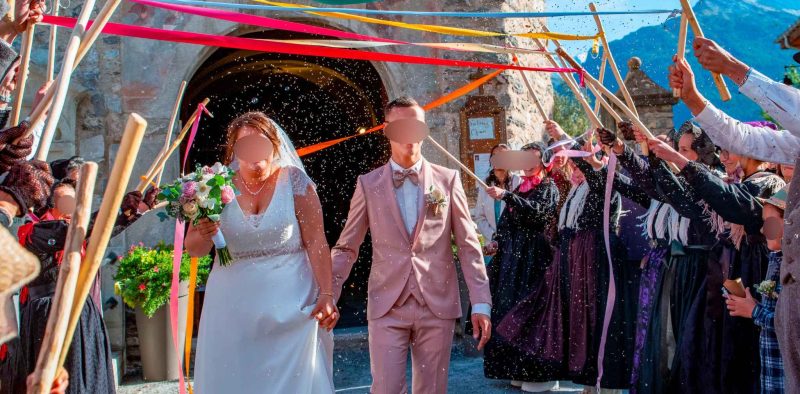 Les mariés sortent de l’église main dans la main, sous une haie d’honneur colorée avec des rubans et des bâtons, entourés d’invités en tenues traditionnelles.