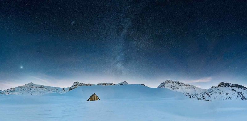 Petite cabane en bois isolée au cœur d’un paysage enneigé sous un ciel nocturne étoilé et la Voie lactée.