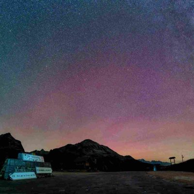Photo nocturne du Col du Galibier avec son panneau en pierre, les silhouettes des montagnes environnantes et un ciel étoilé aux teintes violettes et rosées.