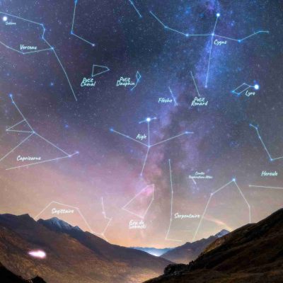 Photographie d’un ciel étoilé nocturne avec les constellations dessinées et nommées, au-dessus d’une vallée de montagne illuminée.