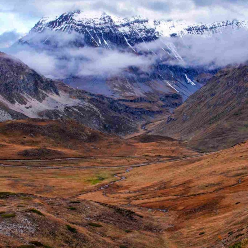 Vallée de montagne aux couleurs automnales avec un sommet enneigé en arrière-plan, partiellement couvert de nuages.