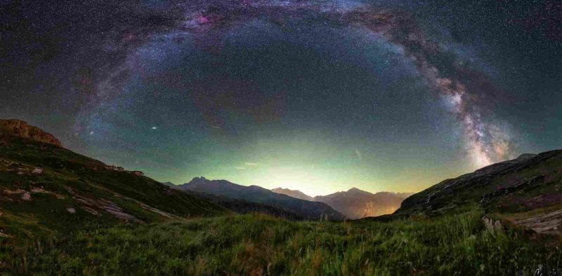 Arc de la Voie lactée illuminant un paysage de montagnes et prairies alpines sous un ciel nocturne dégagé.