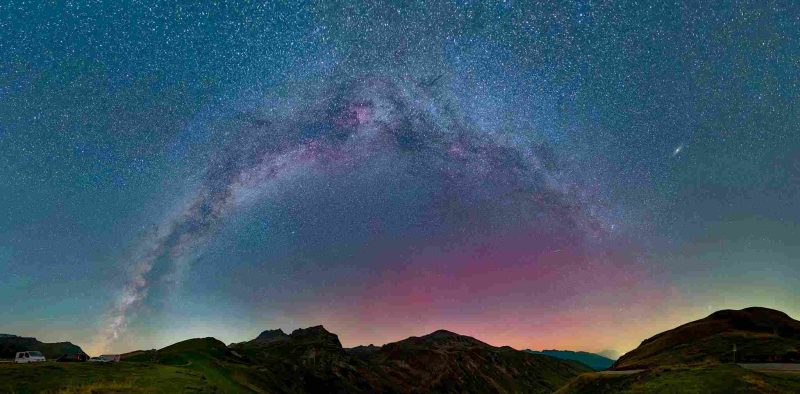 Arc de la Voie lactée traversant le ciel au-dessus de collines et montagnes, sous un ciel étoilé teinté de dégradés de bleu et de rose.