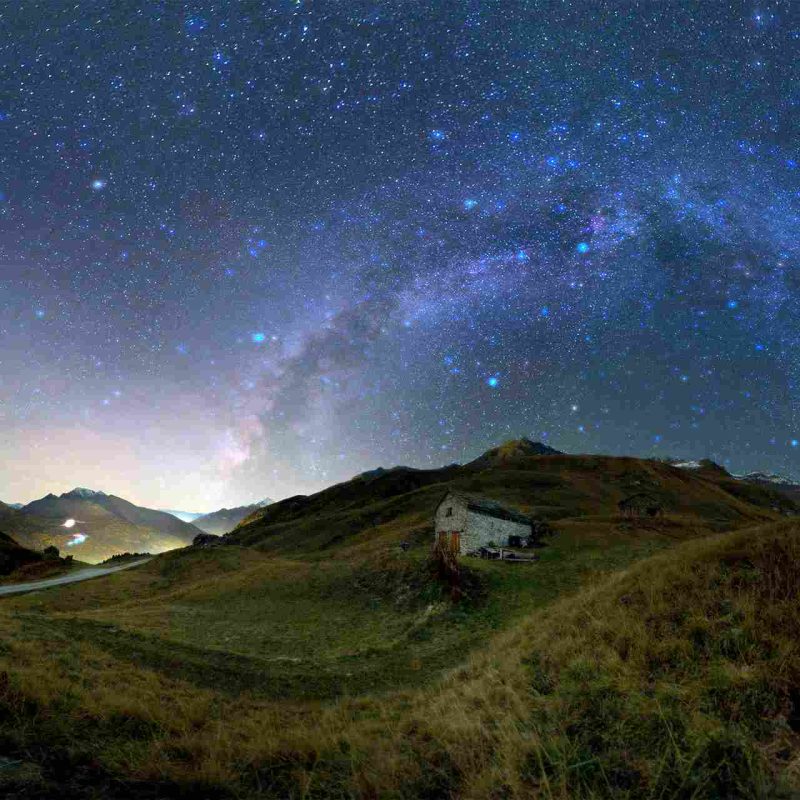Bergerie isolée dans les alpages sous un ciel nocturne scintillant, avec la Voie lactée formant un arc lumineux au-dessus des montagnes.